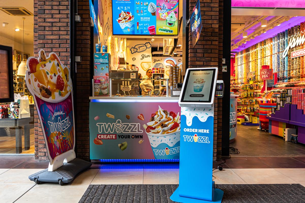 Onesix Kiosk met eigen huisstijl bij de ingang van een ijs- en snoepzaak, met een medewerker achter de balie