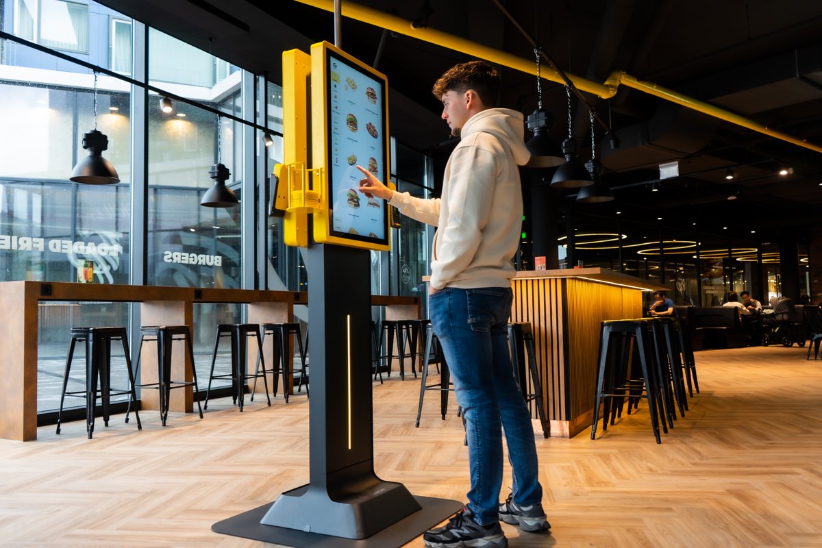 Gast bestelt aan een gele Onesix Kiosk met groot touchscreen in een burgerrestaurant