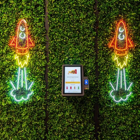 Onesix Kiosk Pro Wall aan een groene plantenwand tussen neon raketten