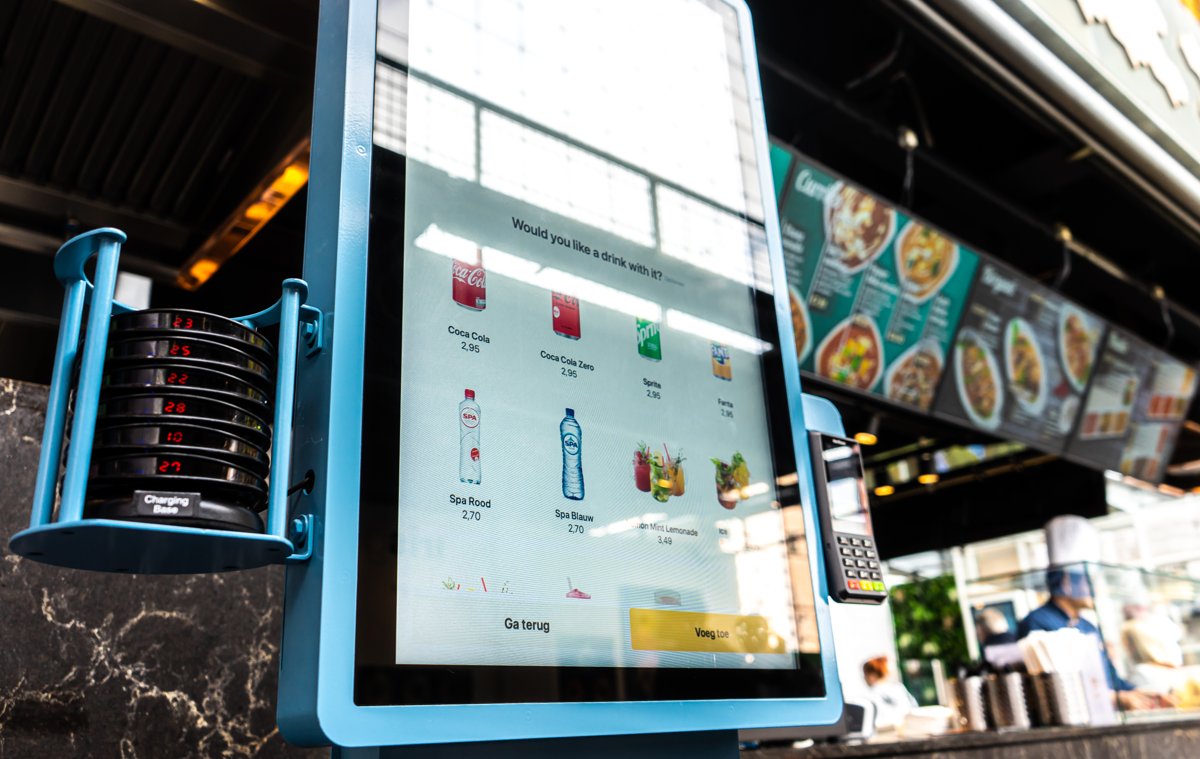 Onesix Kiosk Premium in een drukke markthal, met een Engelse upsell-vraag op het bestelscherm en buzzers op het laadstation