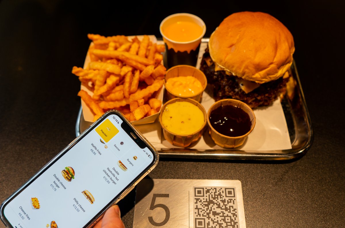Gast bekijkt het digitale menu op de telefoon naast een tafelplaatje met tafelnummer en QR-code, met burger en friet op een dienblad