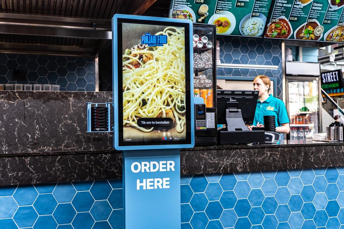 Blauwe Onesix Kiosk met 'Order here' naast de bemande kassa in een Indiaas afhaalrestaurant